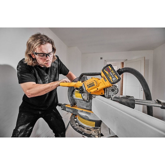 A person operates a DEWALT cordless miter saw (model DCS781B) indoors, making a cut on a long piece of material. The tool is connected to a dust extraction hose. The face of the person is blurred for privacy.