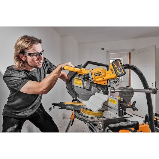 A person is operating a DEWALT DCS781B miter saw, cutting a piece of white molding in a workshop or construction setting. The saw is yellow and black and features a visible dust extraction hose connected to it.