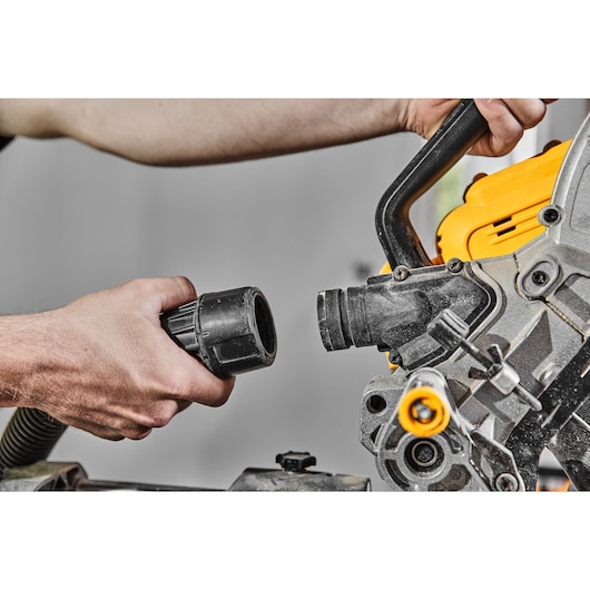 Close-up of a person attaching a vacuum hose to a DEWALT miter saw for dust collection. The hands hold the hose and the dust port of the yellow and black power tool.