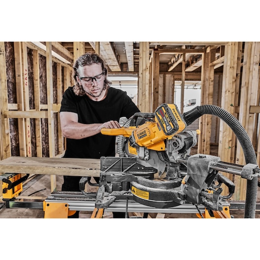 A person using a DEWALT sliding compound miter saw to cut a piece of wood inside a construction site with exposed wooden framing. The saw is equipped with a battery and dust collection hose.