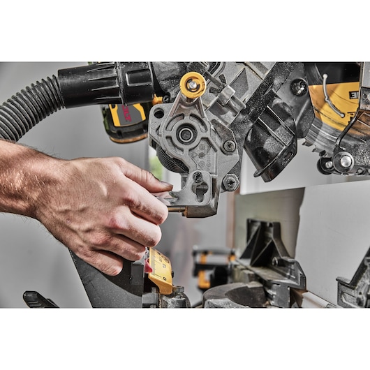 A close-up of a person adjusting the settings on a DEWALT miter saw, model DCS781B, in a workshop setting. The person's hand is operating a lever near the blade and fence, with visible sawdust on the tool.