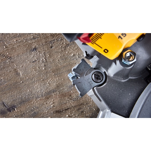 Close-up view of a DEWALT miter saw adjustment mechanism, with a yellow angle indicator and metal components, set on a work surface with wood dust.