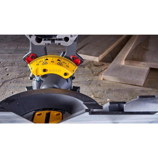 Close-up view of a DEWALT miter saw angle adjustment gauge, showing the yellow scale with degree measurements, mounted on a woodworking surface with wooden boards in the background.