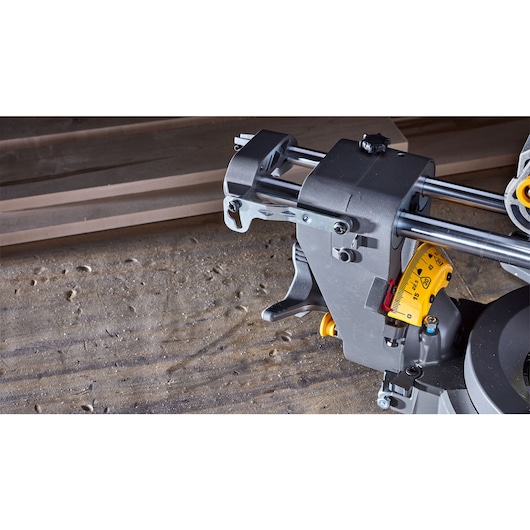 Close-up image of a DEWALT sliding compound miter saw, showing part of the saw's rail mechanism and angle adjustment on a worktable, with wooden boards in the background.