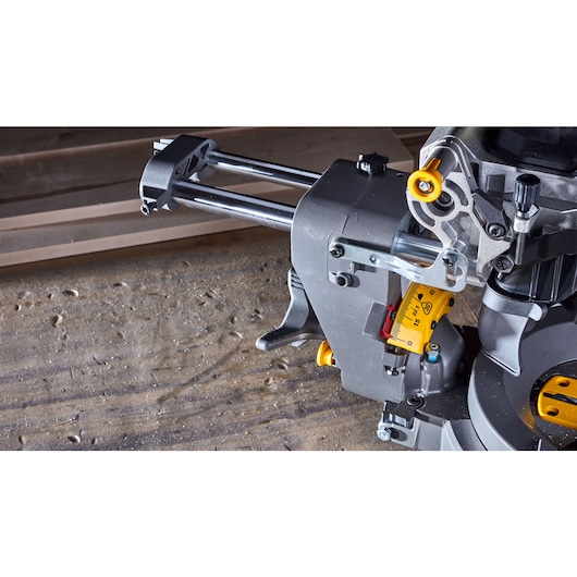 Close-up view of a DEWALT sliding miter saw, showing the metal rails and adjustment mechanisms, set on a dusty workshop surface with wooden boards in the background.