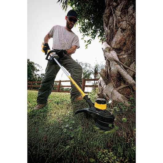 A person using a DEWALT cordless string trimmer to cut grass and weeds next to a large tree in a yard. The trimmer is yellow and black, and the person is wearing gloves and work clothes.