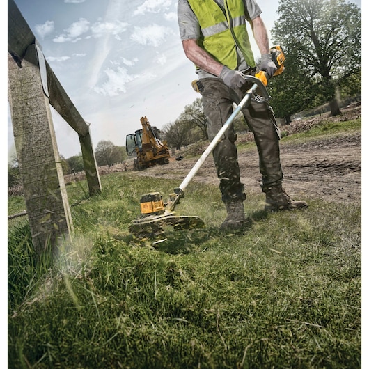 A person wearing a safety vest and gloves is using a DEWALT DCST920B string trimmer to cut grass along a fence outdoors. There is construction equipment in the background and the area is on a dirt path surrounded by greenery.