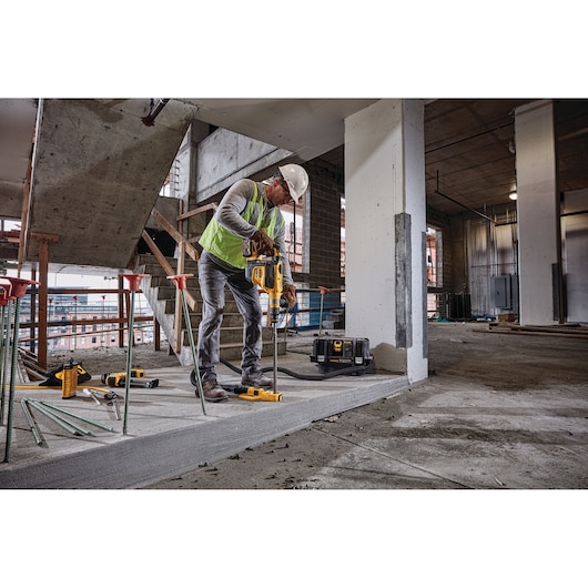 A construction worker wearing a hard hat and safety vest uses a DEWALT power tool to drill into a concrete floor at an indoor construction site. Nearby, multiple DEWALT tools and a DEWALT vacuum are visible.