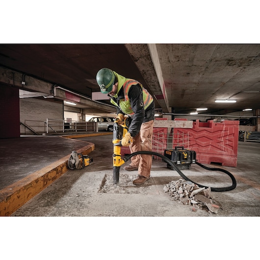 A construction worker is using a DEWALT cordless dust extractor (model DCV585B) connected to a power tool to remove debris and dust while working on a concrete floor in a parking garage.