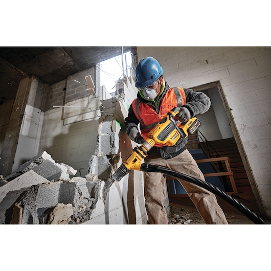 A construction worker wearing a blue hard hat and orange safety vest uses a DEWALT power tool to demolish a concrete wall. Rubble and debris are scattered on the ground, and the worker is using a vacuum hose attachment connected to the tool for dust control.