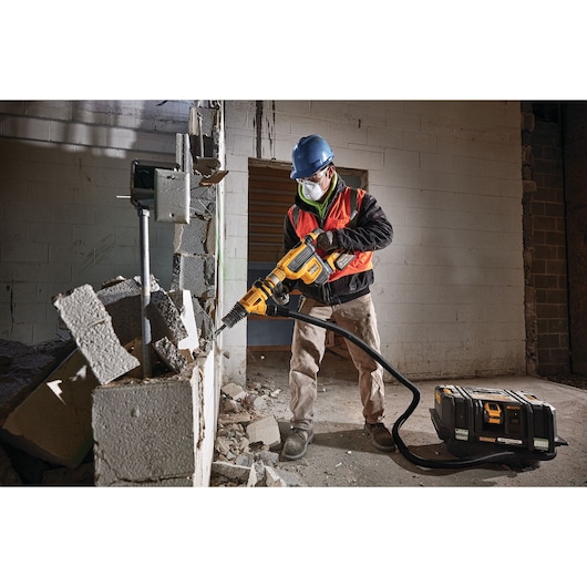 A construction worker wearing safety gear is using a DEWALT DCV585B dust extractor connected to a power tool to break down a concrete block wall. Debris is scattered around the work area.
