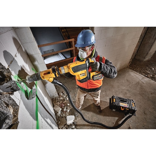 A construction worker wearing a blue hard hat and orange safety vest is using a DEWALT power tool connected to a vacuum system to drill into a concrete wall. Debris and dust are visible on the ground, and a DEWALT toolbox is placed nearby.