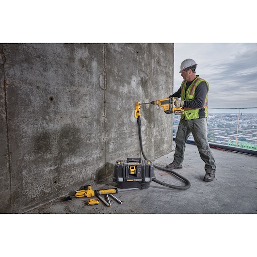 A construction worker wearing safety gear uses a DEWALT cordless vacuum system (DCV585B) attached to a drill, working on a concrete wall. The DEWALT vacuum and additional accessories are placed on the ground.
