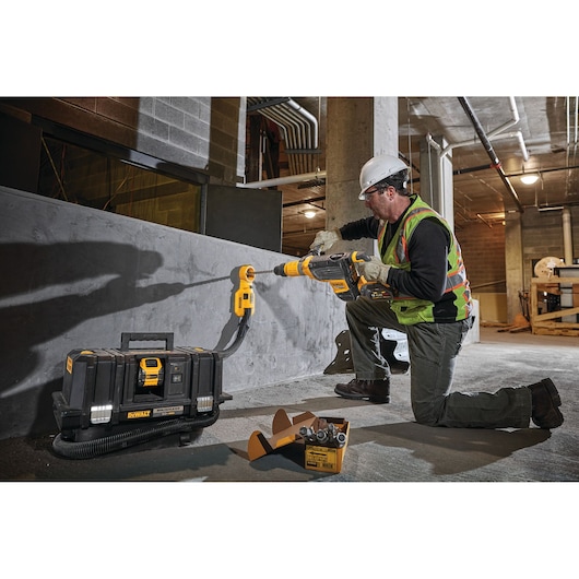 A construction worker wearing a safety helmet and high-visibility vest is using a DeWalt power tool to drill into a concrete wall. Next to him is a DeWalt portable dust extractor DCV585B and other tool accessories in a construction site environment.