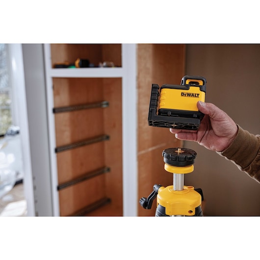 A person is holding a DEWALT laser level device, about to attach it onto a tripod. The scene is set indoors near a cabinet under construction.