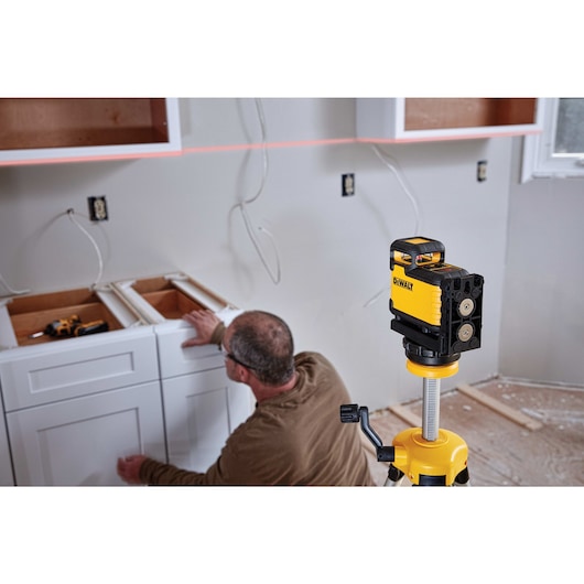 A DEWALT laser level device is set up on a tripod in a room under renovation, helping align cabinets being installed by a worker.