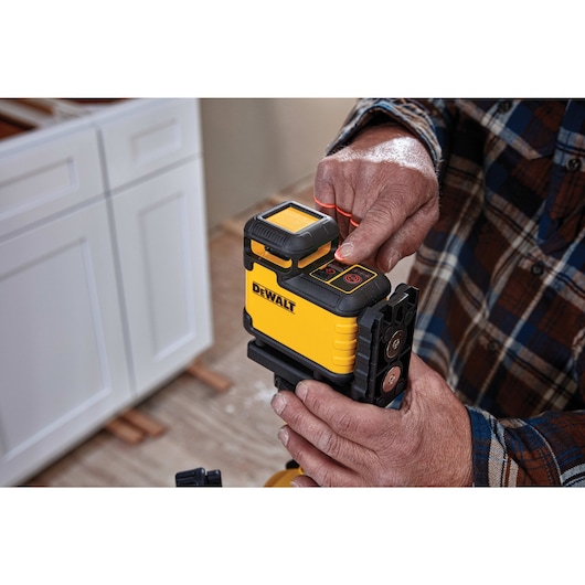 Close-up of a person operating a yellow DEWALT laser level device, which is positioned on a mounting bracket. The person's finger is pressing a button on the top panel. Nearby, a kitchen cabinet and a plaid shirt sleeve are visible.