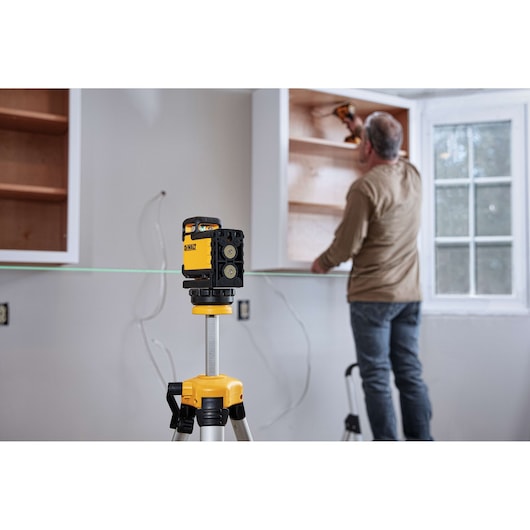 A DEWALT laser level tool is set up on a tripod, projecting a green laser line across a room where a person is installing or adjusting shelves in a cabinet against the wall.