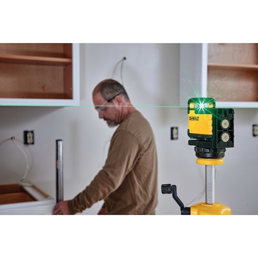 A DEWALT green line laser level is set up on a tripod in a room under construction, emitting a green laser across the space. A person with a blurred face is working in the background near cabinets and wall outlets.
