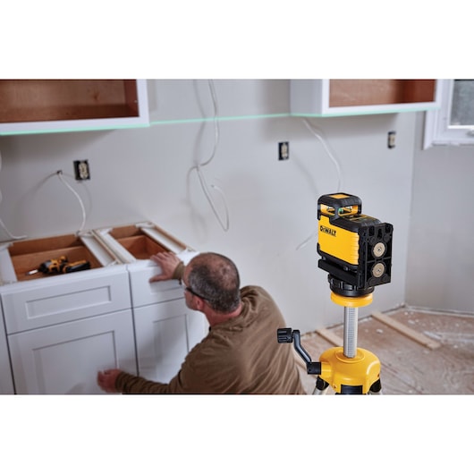 A DEWALT laser level device is mounted on a tripod in the foreground, with a person installing white cabinets in a kitchen under construction. The walls have exposed wires and there are open cabinet frames and tools visible.