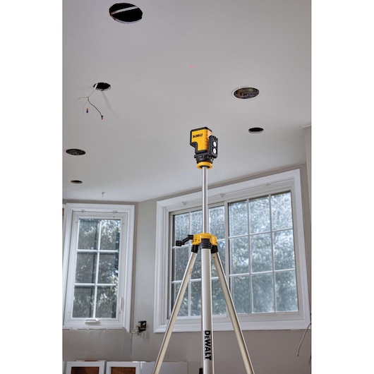 A DEWALT laser level device mounted on a tripod, positioned indoors in a room with large windows, used for ceiling installation work.