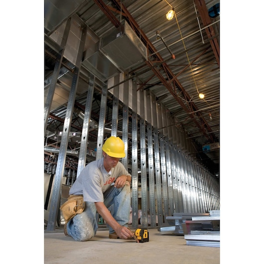 A worker wearing a yellow hard hat is kneeling on the floor of a construction site, using a DEWALT DW083K laser level. Steel wall studs and metal framing are visible in the background, with exposed ceiling and lighting overhead.
