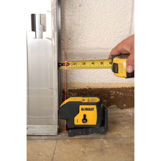 A DEWALT DW083K laser level placed on the floor next to a metal frame, projecting a red vertical laser line. A hand is holding a yellow tape measure against the wall, measuring the distance from the laser line to the metal frame.