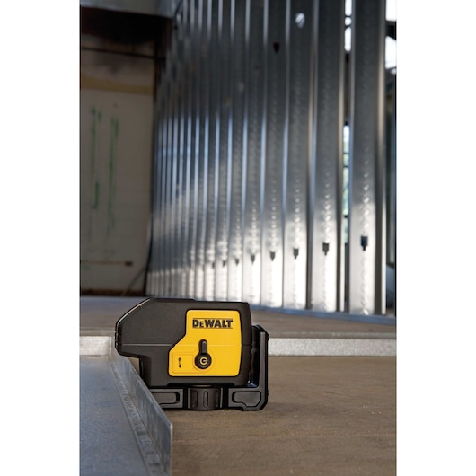 A DEWALT laser level (model DW083K) is positioned on a construction site floor, next to metal wall framing studs. The yellow and black device is designed for precise alignment and leveling tasks.