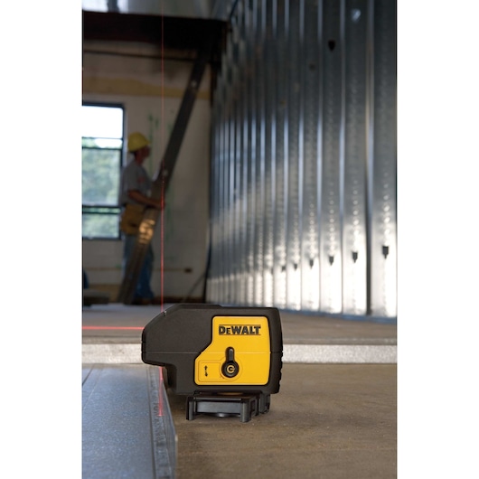 A DEWALT DW083K laser level tool is positioned on the floor in an industrial setting, projecting a red vertical line onto metal studs. In the background, a person wearing a hard hat is working near a window.