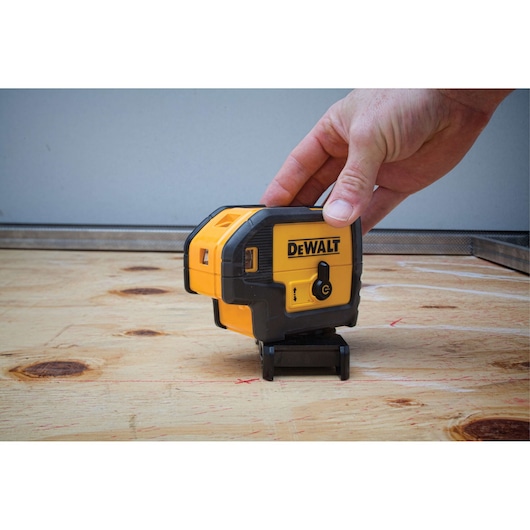 A person’s hand is adjusting a DEWALT DW085K line laser level on a wooden surface. The laser level is yellow and black and is used for precise alignment in construction or photography setups.