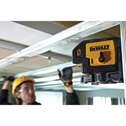 A DEWALT laser level device mounted on a metal frame, projecting a red laser line across the ceiling. A person wearing a yellow hard hat and work gloves is adjusting overhead components in the background.