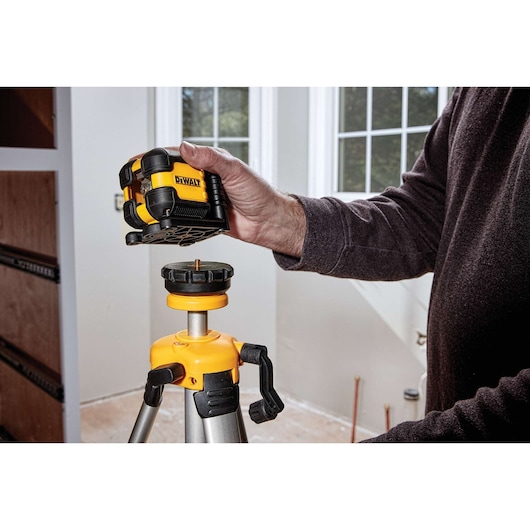A person is placing a DEWALT DW08802 laser level onto a tripod inside a room with windows.