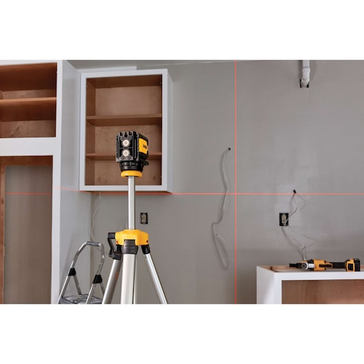 A DEWALT laser level on a tripod projects red horizontal and vertical lines onto a wall in a room under construction. Shelving, wiring, and tools are visible in the workspace.