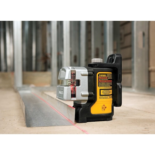 A DEWALT DW089K self-leveling line laser placed on a construction site floor, projecting a visible red laser line onto a metal track for precise alignment during building framing.