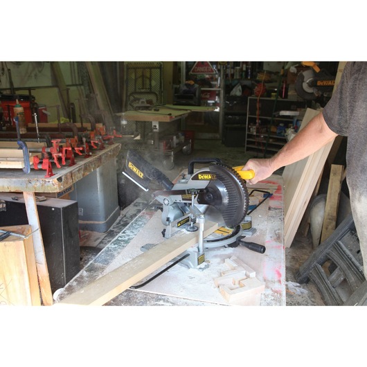 A person operating a DEWALT miter saw, model DW714, to cut a piece of wood in a workshop setting. Sawdust and woodworking tools are visible on and around the workbench.