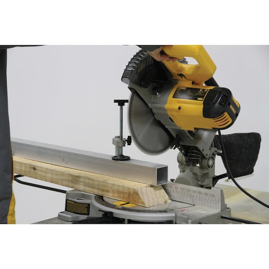 A DEWALT DW714 miter saw being used to cut a piece of metal and a wooden beam. The saw is yellow and black, and the metal and wood are secured on the saw base.