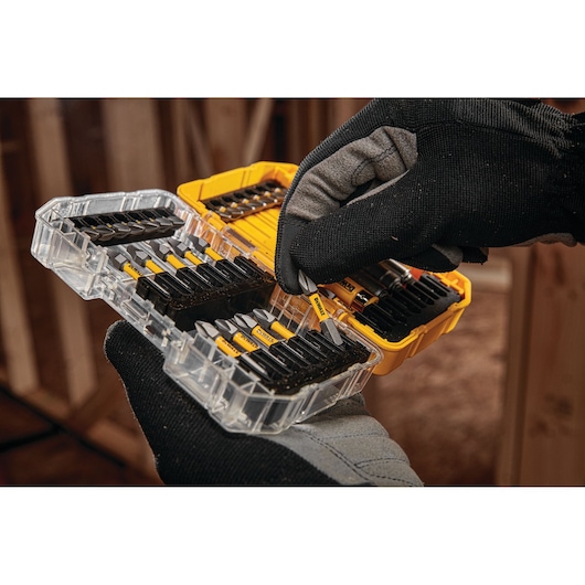 A gloved hand is selecting a screwdriver bit from a transparent plastic case containing various DEWALT branded bits. The case is yellow and opened, showing several rows of bits organized neatly inside.