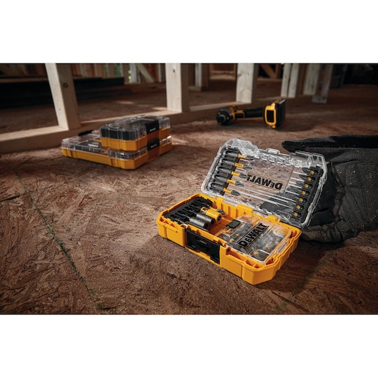 A DEWALT yellow and black tool kit is open on a construction site floor, displaying various drill bits and accessories inside a clear plastic lid. Another closed tool kit and a cordless tool are visible in the background. A work glove rests beside the open kit.