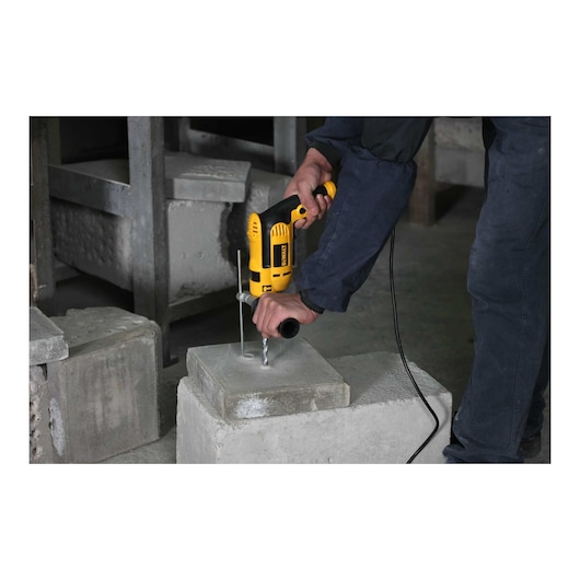 A person is using a yellow and black DEWALT electric drill to drill into a concrete block, with construction materials visible in the background.