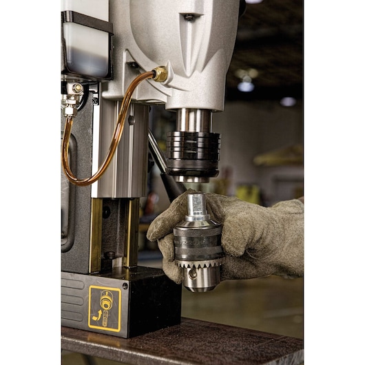 A close-up of a person wearing a work glove attaching a drill chuck to a DEWALT magnetic drill press. The image shows the drill's metal components, a lubricant hose, and an instructional sticker on the base.