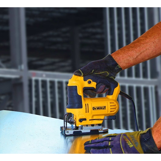 A person wearing work gloves is using a yellow and black DEWALT jigsaw to cut a sheet of metal.