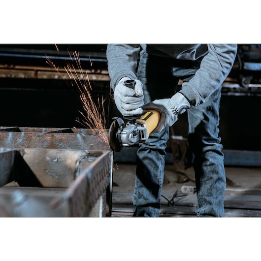 A person wearing protective gloves uses a yellow DEWALT angle grinder (model DWE4010) to cut or grind a piece of metal, producing sparks in an industrial workshop setting.
