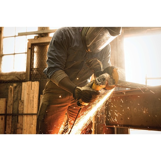 A person wearing protective gear uses a DEWALT angle grinder, producing sparks as they cut or grind a piece of metal in an industrial workshop setting.