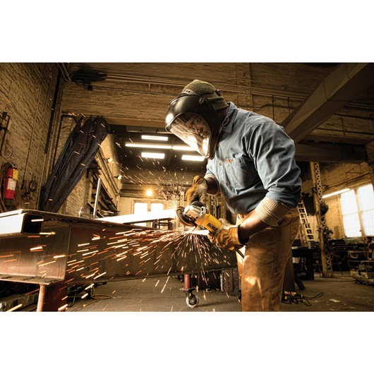 A worker wearing protective gear, including a face shield and gloves, uses a DEWALT angle grinder to cut metal in an industrial workshop, with sparks flying from the grinding process.