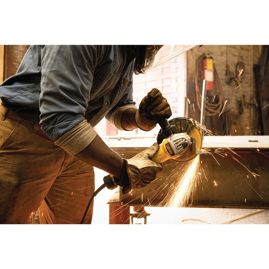 A person wearing protective gloves and a face shield is using a DEWALT angle grinder, generating sparks while working on a metal surface in an industrial workshop.