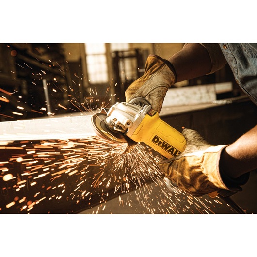 A person wearing protective gloves is using a DEWALT angle grinder to cut metal, with sparks flying in a workshop environment.