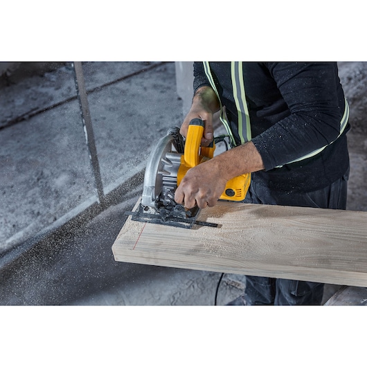 A person using a yellow DEWALT circular saw (model DWE5615) to cut a wooden plank, with sawdust and safety gear visible.