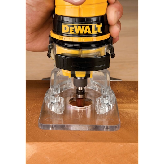 Close-up of a DEWALT handheld router being used to cut or shape wood on a work surface. The router is held by a person's hand and has a transparent base for visibility.