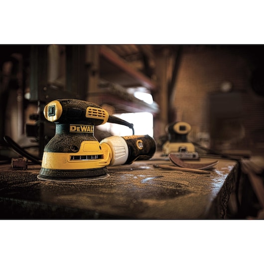 A DEWALT DWE6421 yellow and black electric orbital sander is placed on a dusty workbench in a workshop. There are additional sanding tools and sandpaper nearby in the background.