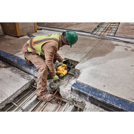 A person wearing safety gear and a green helmet uses a DEWALT DWH205DH tool to cut into a concrete surface at a construction site. Rebar is visible underneath the concrete.